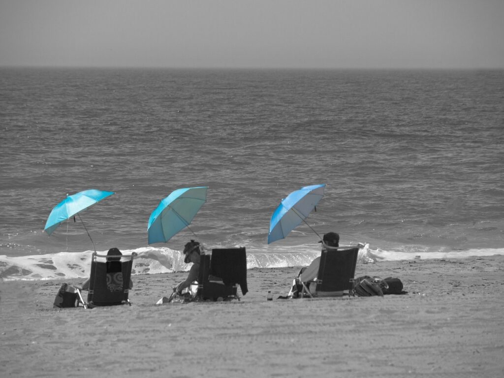 Trois personnes sur la plage, en face de la mer, sur des transats et sous des parasols bleus.