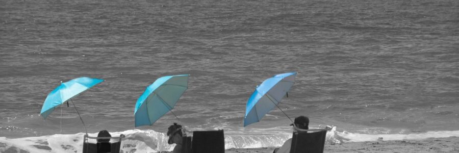 Trois personnes sur la plage, en face de la mer, sur des transats et sous des parasols bleus.